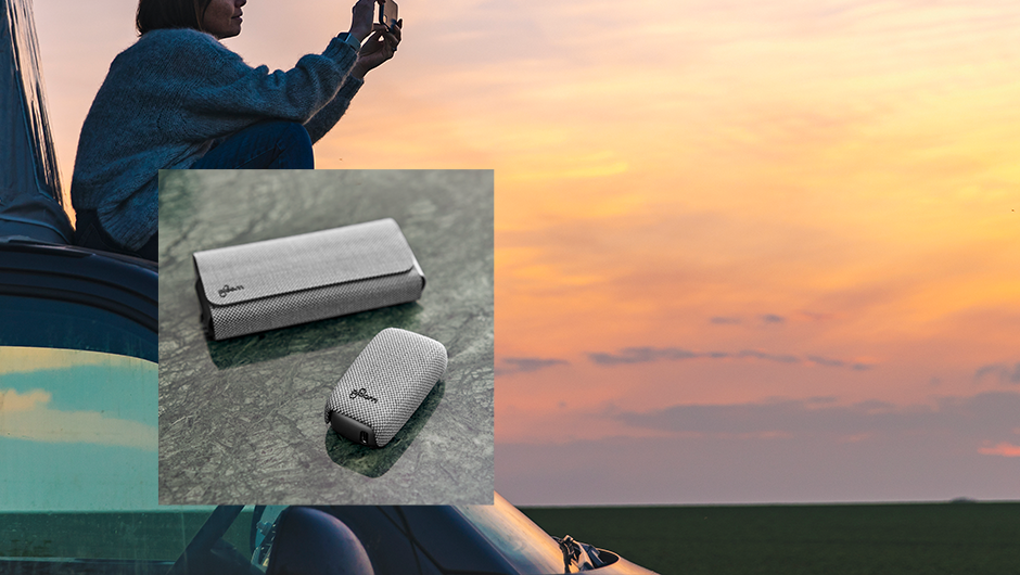Žena sedící na automobilu fotící západ slunce, v popředí zařízení Ploom X Advanced s textilním krytem a pouzdrem