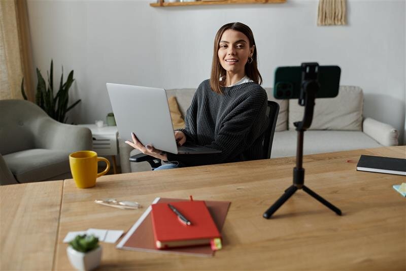 Eine Frau sitzt an einem Tisch mit dem Laptop auf dem Arm. Sie schaut in ein Smartphone auf einem Stativ, weil sie sich selber filmt.