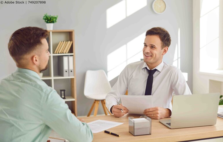 Zwei Männer die in einem Büro am Tisch sitzen und etwas besprechen.