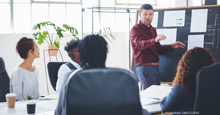 Ein Mann präsentiert im Büro vor einem Team an einem Flipchart und erklärt Strategien und Diagramme während die Zuhörer aufmerksam folgen.