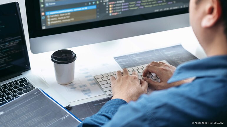 Eine Person tippt konzentriert an einem Computer, während auf dem Bildschirm Programmcode angezeigt wird und Unterlagen auf dem Schreibtisch liegen.