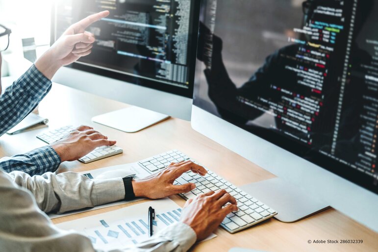 Zwei Personen arbeiten gemeinsam an Softwarecode und digitalen Prozessen an großen Bildschirmen in einer modernen Büroumgebung.