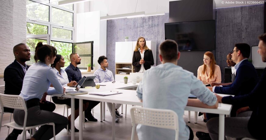 Many people sitting at in a meeting room with a person talking in front of them