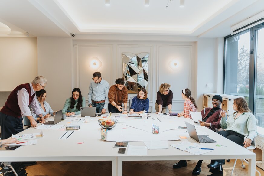 10 Menschen sitzen an einem Tisch mit Papier und Laptops und unterhalten sich angeregt mit einander.