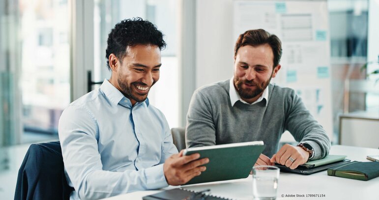 Zwei Kollegen sitzen gemeinsam am Tisch, schauen auf ein Tablet und tauschen sich in entspannter, konzentrierter Atmosphäre über Inhalte oder Ergebnisse aus.