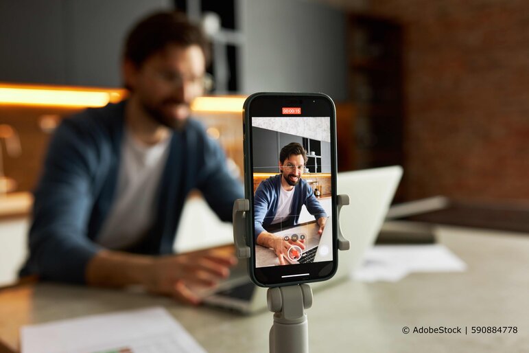 Ein Mann erstellt mit seinem Smartphone ein Social-Media-Video, während er an einem Laptop arbeitet.