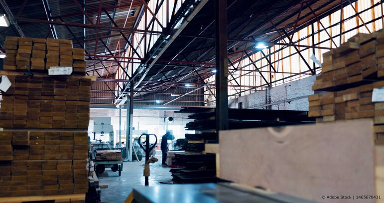 Industriehalle mit gestapelten Holzmaterialien, Produktionsmaschinen und einem Mitarbeiter in einer weitläufigen Fertigungsumgebung.