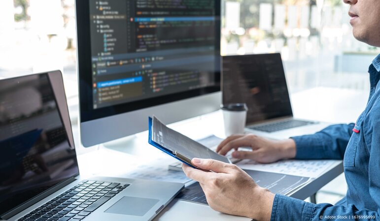 Eine Person arbeitet am Computer mit sichtbarem Programmcode und überprüft parallel digitale Unterlagen an einem modernen Arbeitsplatz.