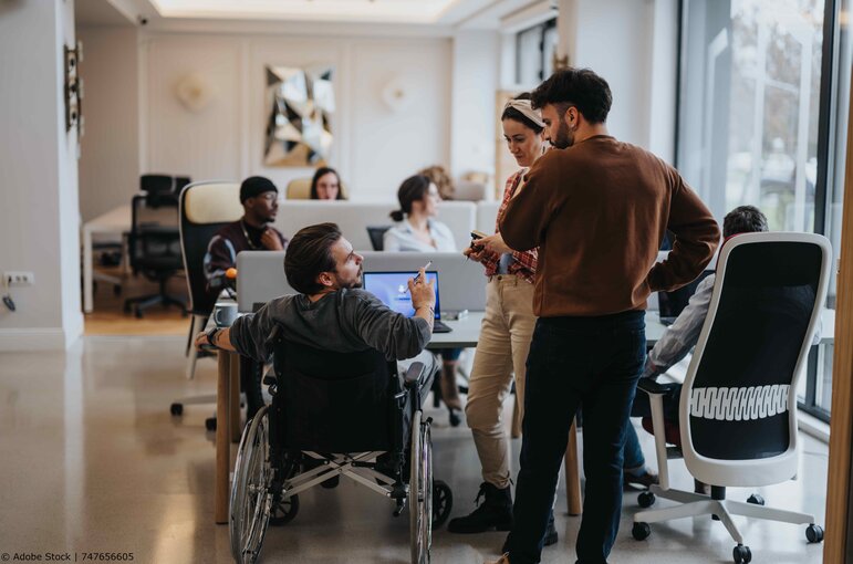 Ein Team von drei Personen, darunter ein Mann, der im Rollstuhl sitzt, unterhalten sich im Büro miteinander