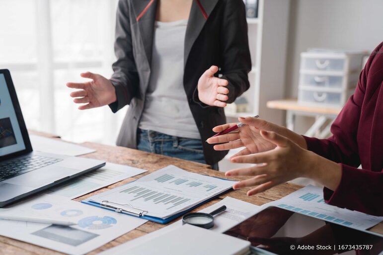 Geschäftspersonen stehen an einem Tisch und diskutieren anhand eines Laptops und ausgedruckter Diagramme.