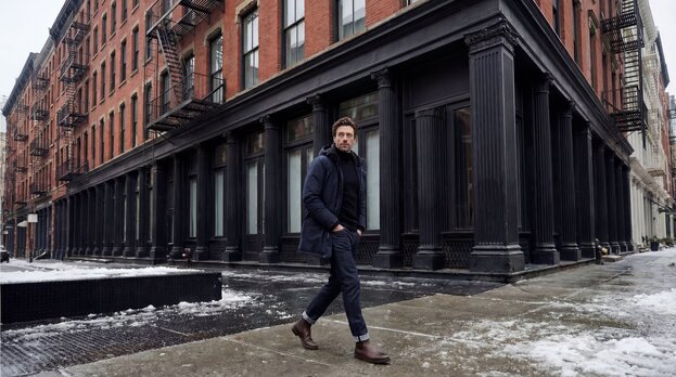A man walking in a snow-covered sidewalk wearing a navy parka and waxed chelsea boots