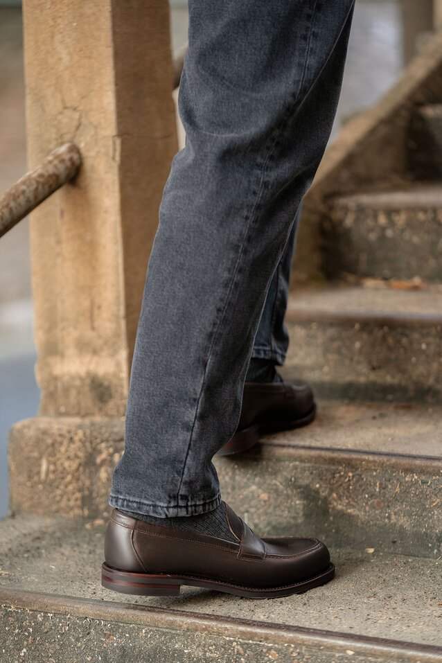 Person wearing a pair of grey jeans and a pair of brown waxy calf leather loafers on a concrete stair.