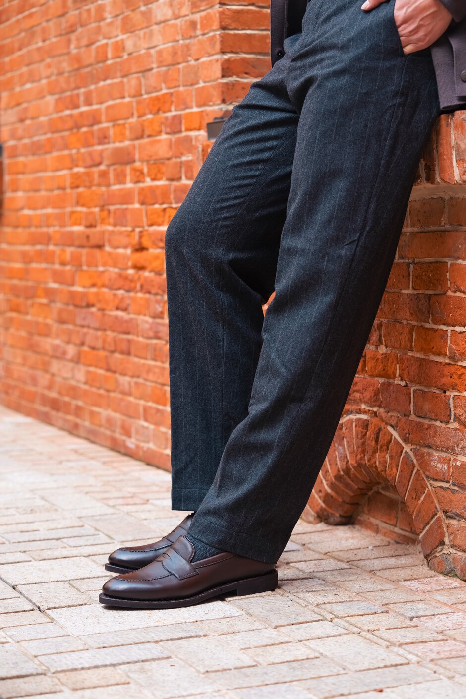 Person in dark dress pants wearing brown leather loafers, standing near a rustic stone wall.