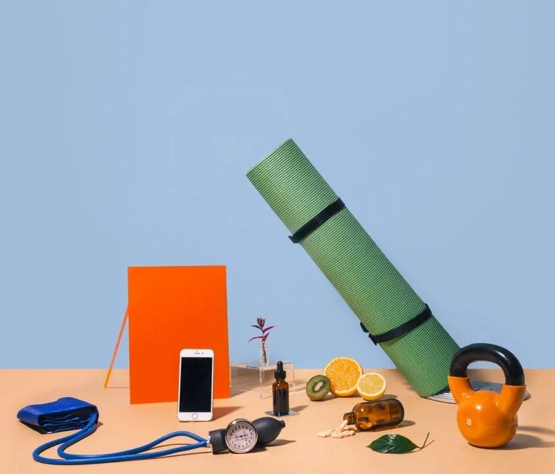 Health and wellness items on a flat surface, including a yoga mat, kettlebell, phone, blood pressure monitor, fruits, supplements, and a dropper bottle.