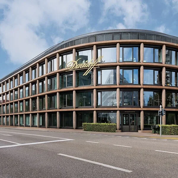 Ein modernes Glasgebäude mit einer Außenfassade, die an Säulen in Form von Zigarren erinnert. Dies ist das Maison Davidoff Gebäude, das als Hauptsitz in Basel dient.