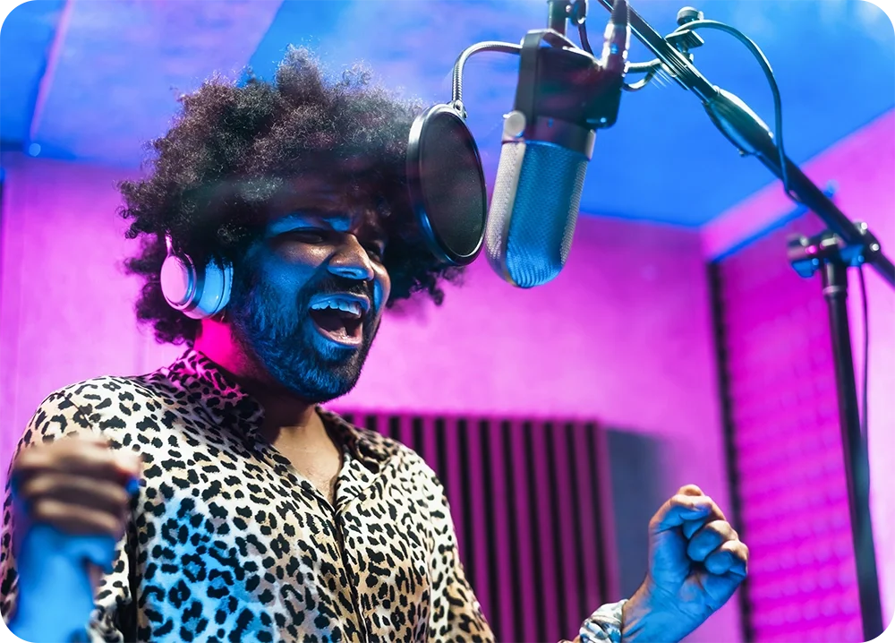 A man in a vocal booth with hands up singing powerfully into a microphone.