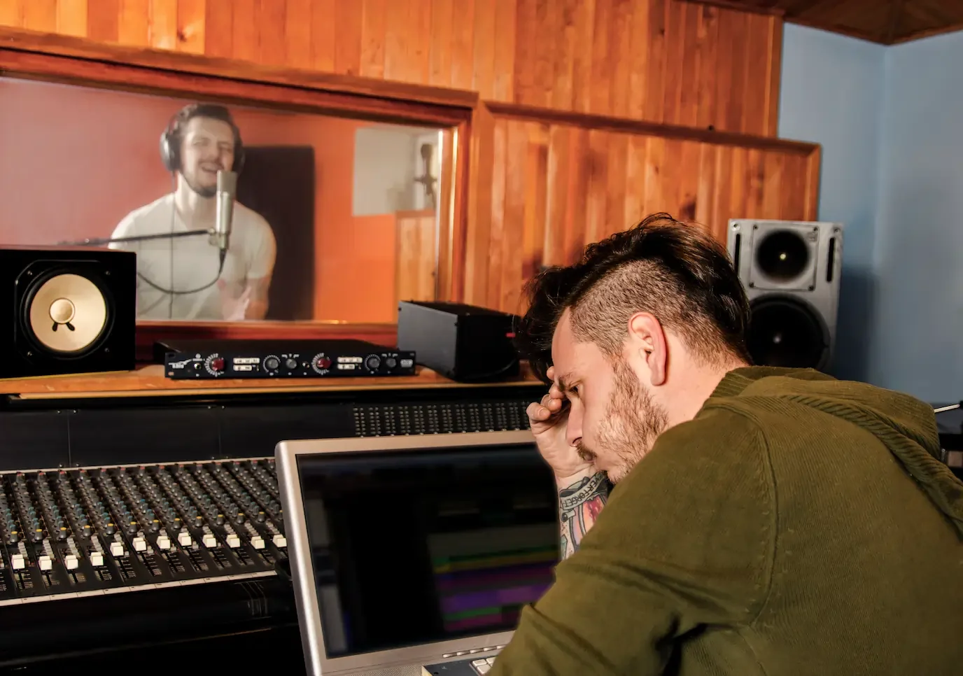 A man with his head in his hand recording another man singing into a microphone in a recording booth