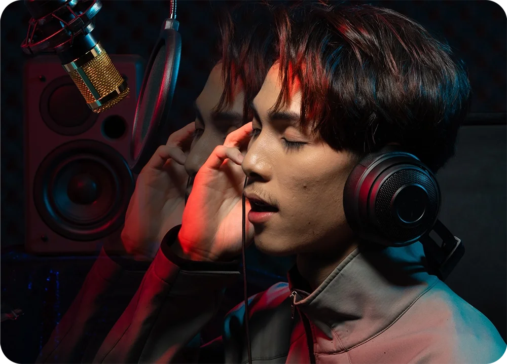 A person in a recording studio wears large headphones and holds one ear cup while singing into a suspended microphone. Red and blue accent lighting illuminates the scene, with a speaker visible in the background and a faint double-exposure effect creating a mirrored profile.
