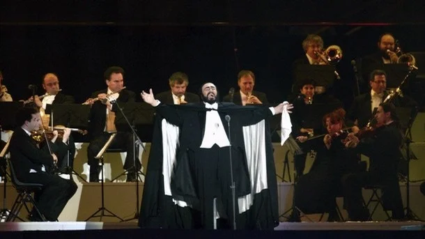 Luciano Pavorotti on stage with his arms stretched wide with an orchestra behind him