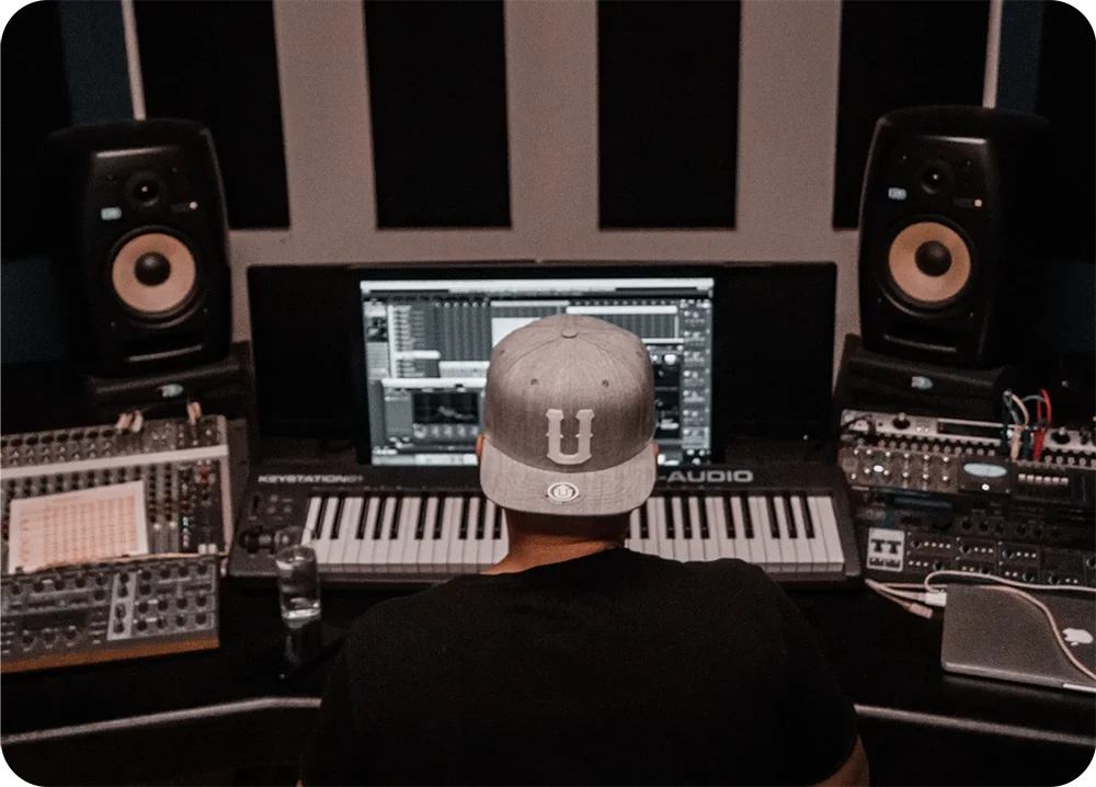 Person seated at a music production workstation with a keyboard, studio monitors, and computer screen.