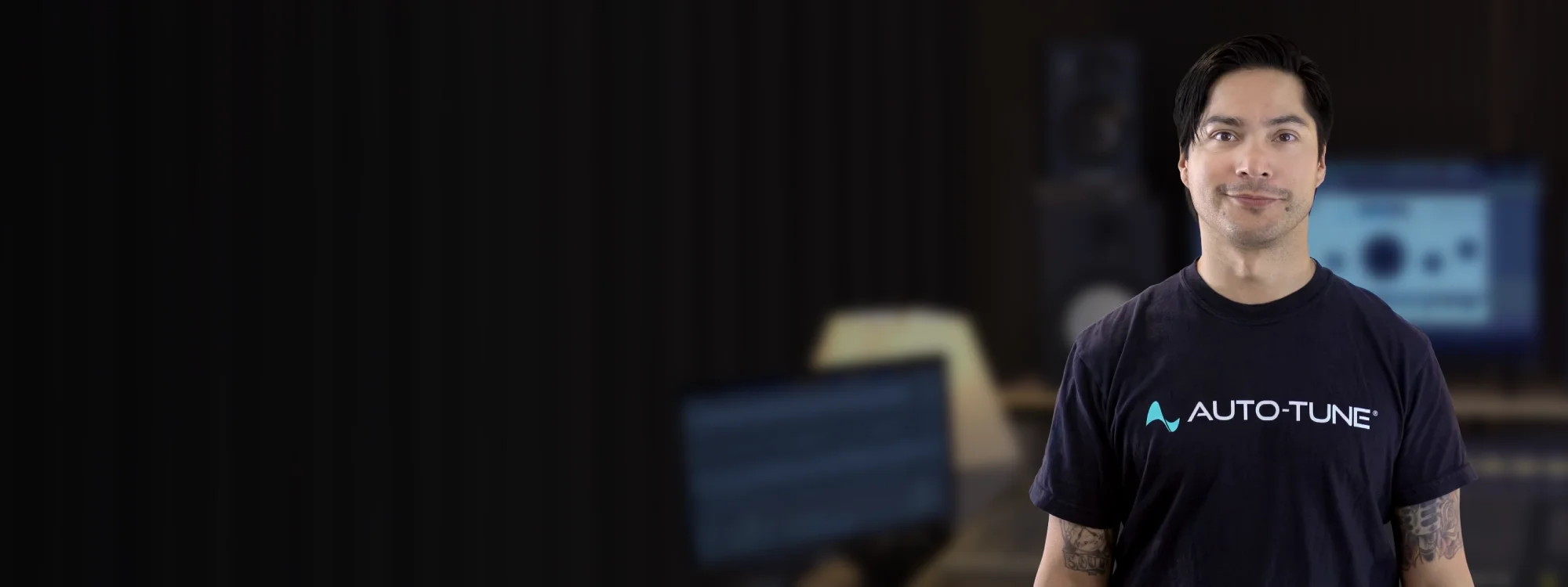 A man in a black AutoTune t-shirt standing in front of a blurred home studio setup