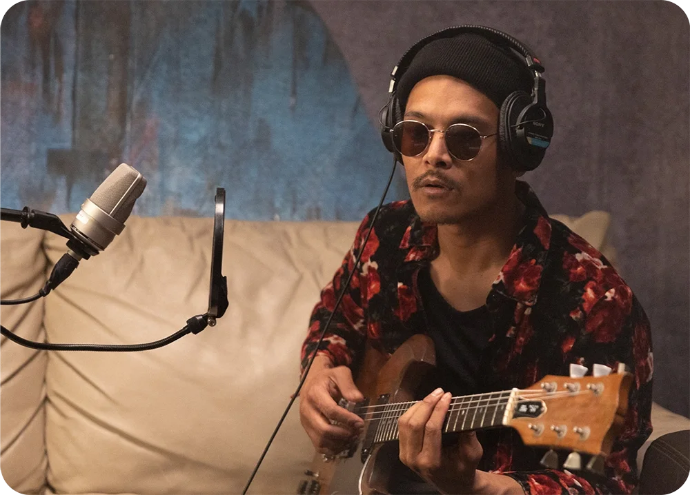 A man wearing headphones sitting on a couch playing guitar while singing into a microphone