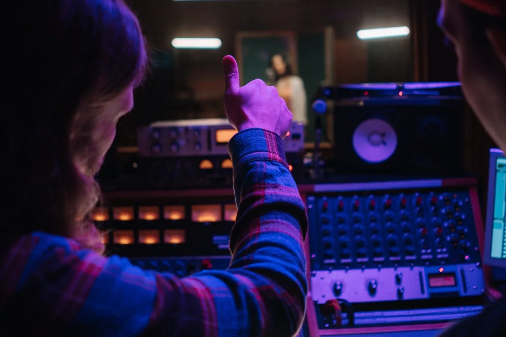 A man sitting in front of a mixer giving a thumbs up to a singer in the booth