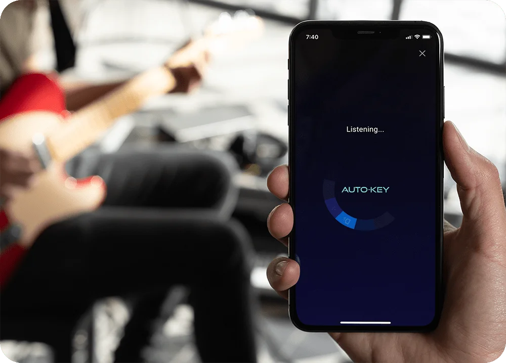 A hand holding an iPhone with AutoKey Mobile listening to a person playing guitar in background