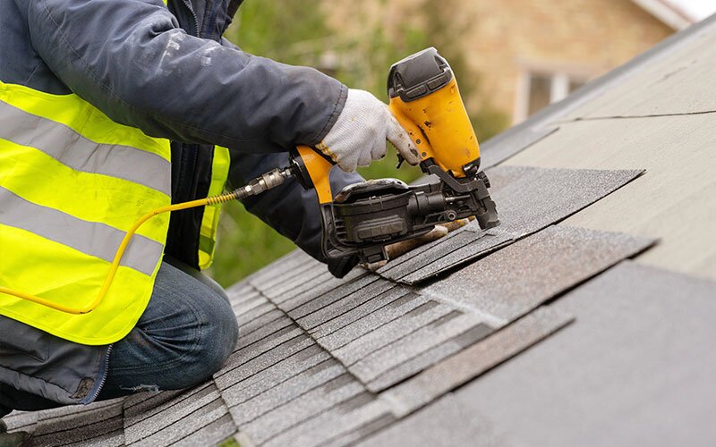 A roofer is installing asphalt shingles on a roof using a nailer gun.