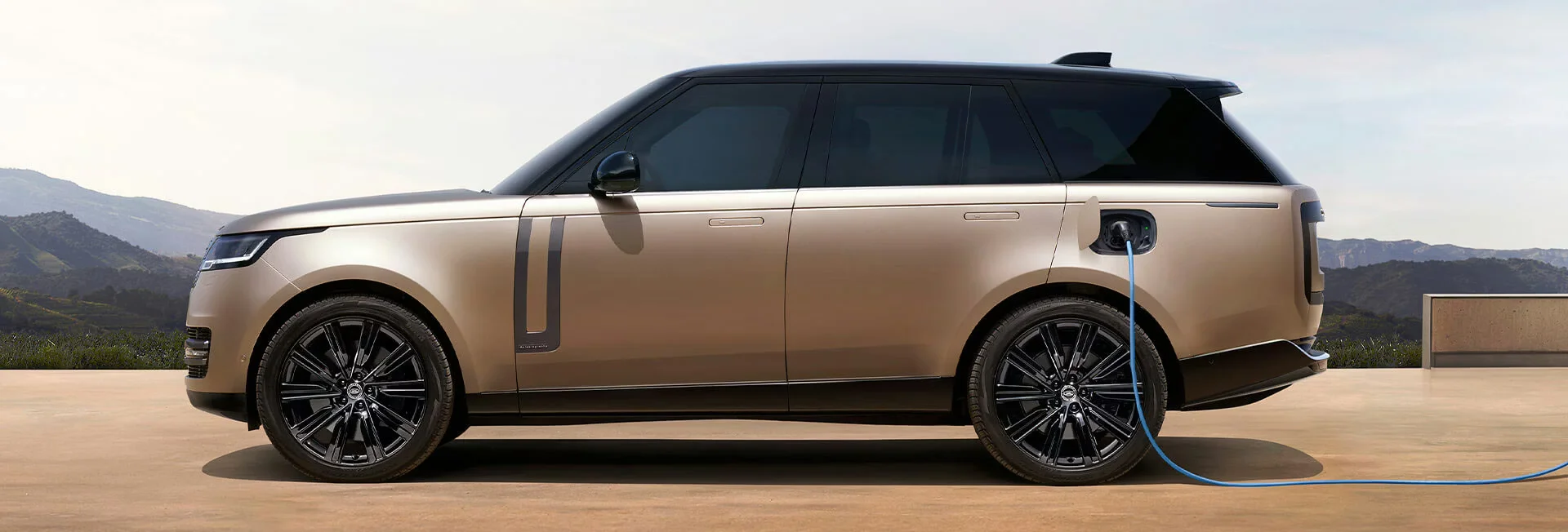 Blauer Elektro‑SUV fährt bei Sonnenuntergang auf einer Bergstraße, im Hintergrund schneebedeckte Gipfel und warmer Himmel.