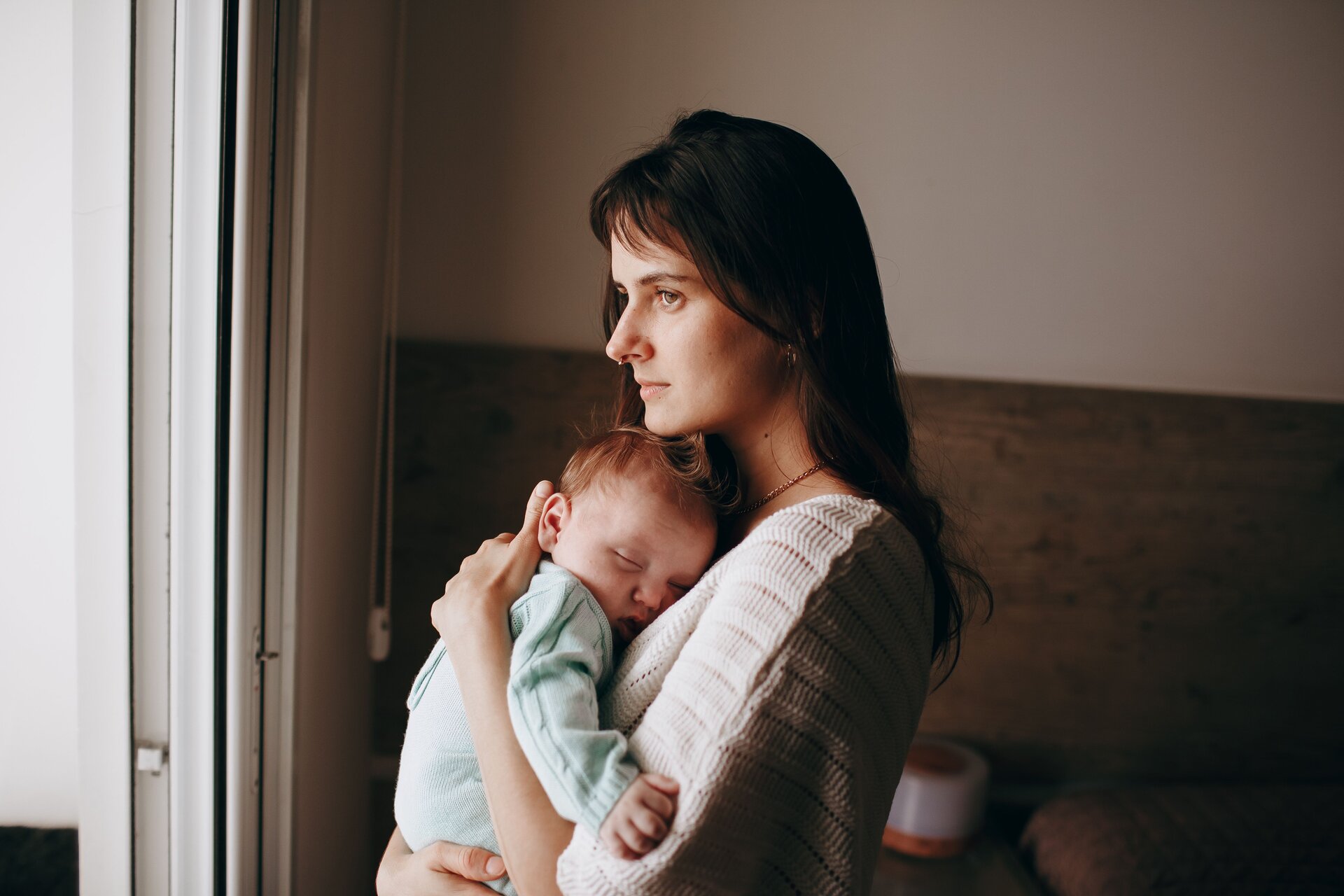 A woman stands and looks off to the left while holding a baby to her chest
