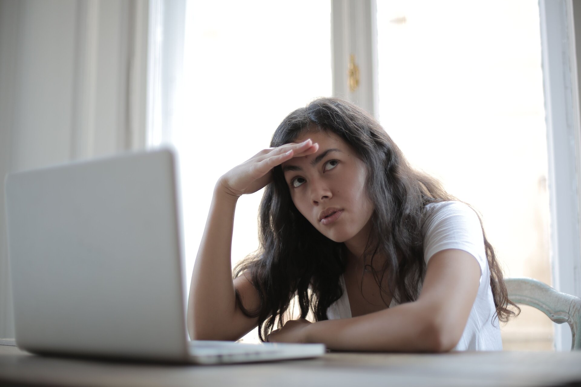 A young woman sits in front of a laptop and looks off tot he right with her hand on her forehead