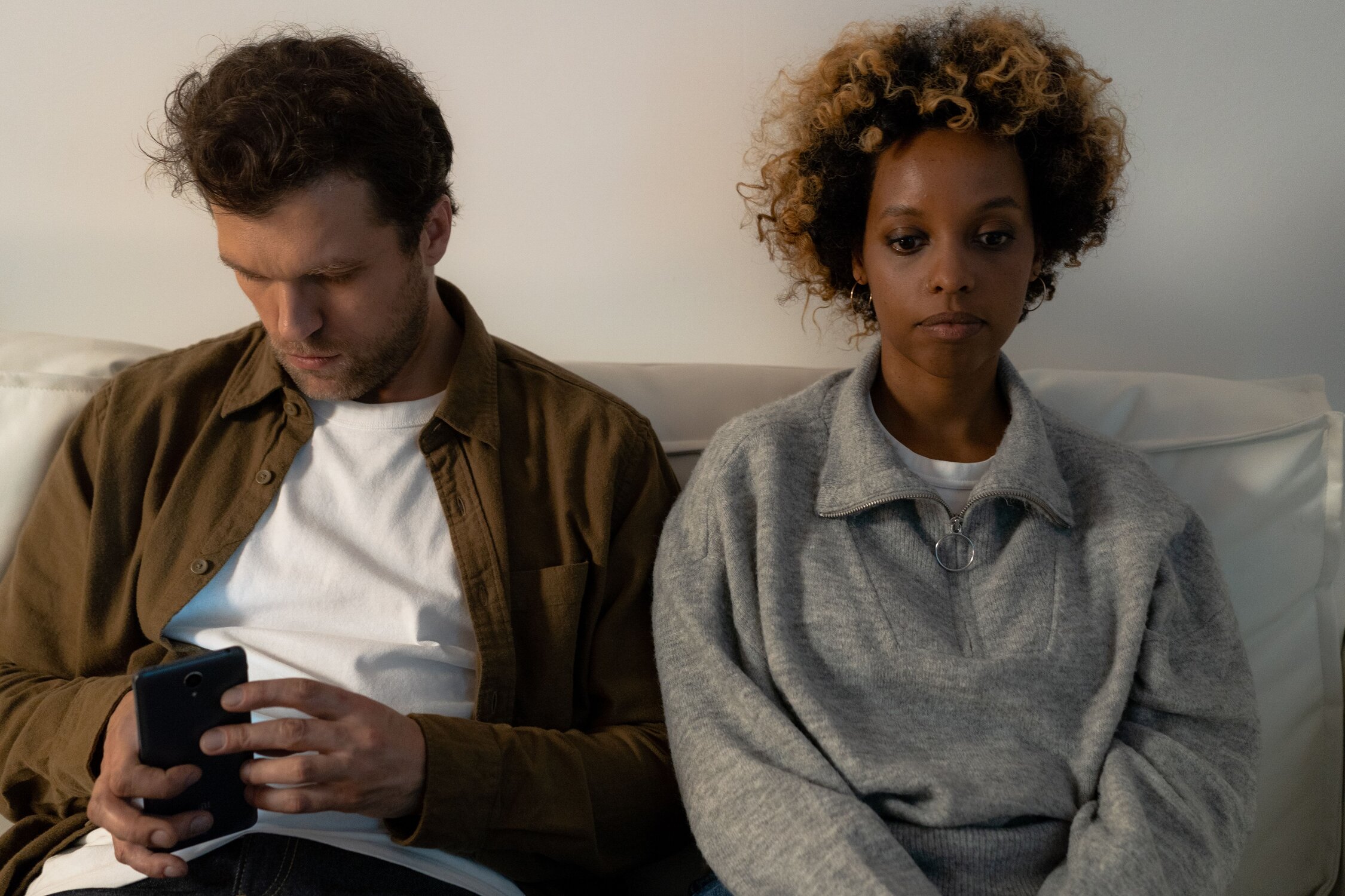 A woman sits on the couch staring into the distance while a man sits next to her and looks at his phone