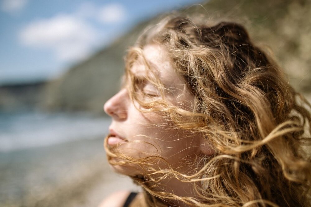 Person at the beach with their eyes closed while the wind blows their hair back