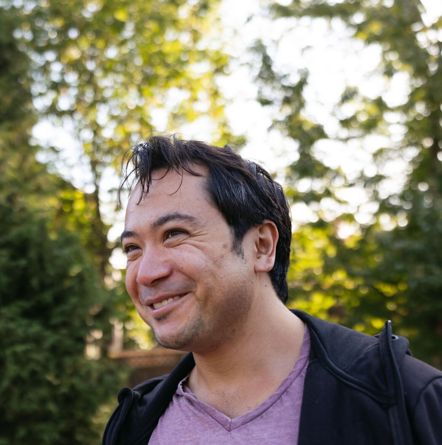 A smiling man outdoors with trees and sunlight in the background.