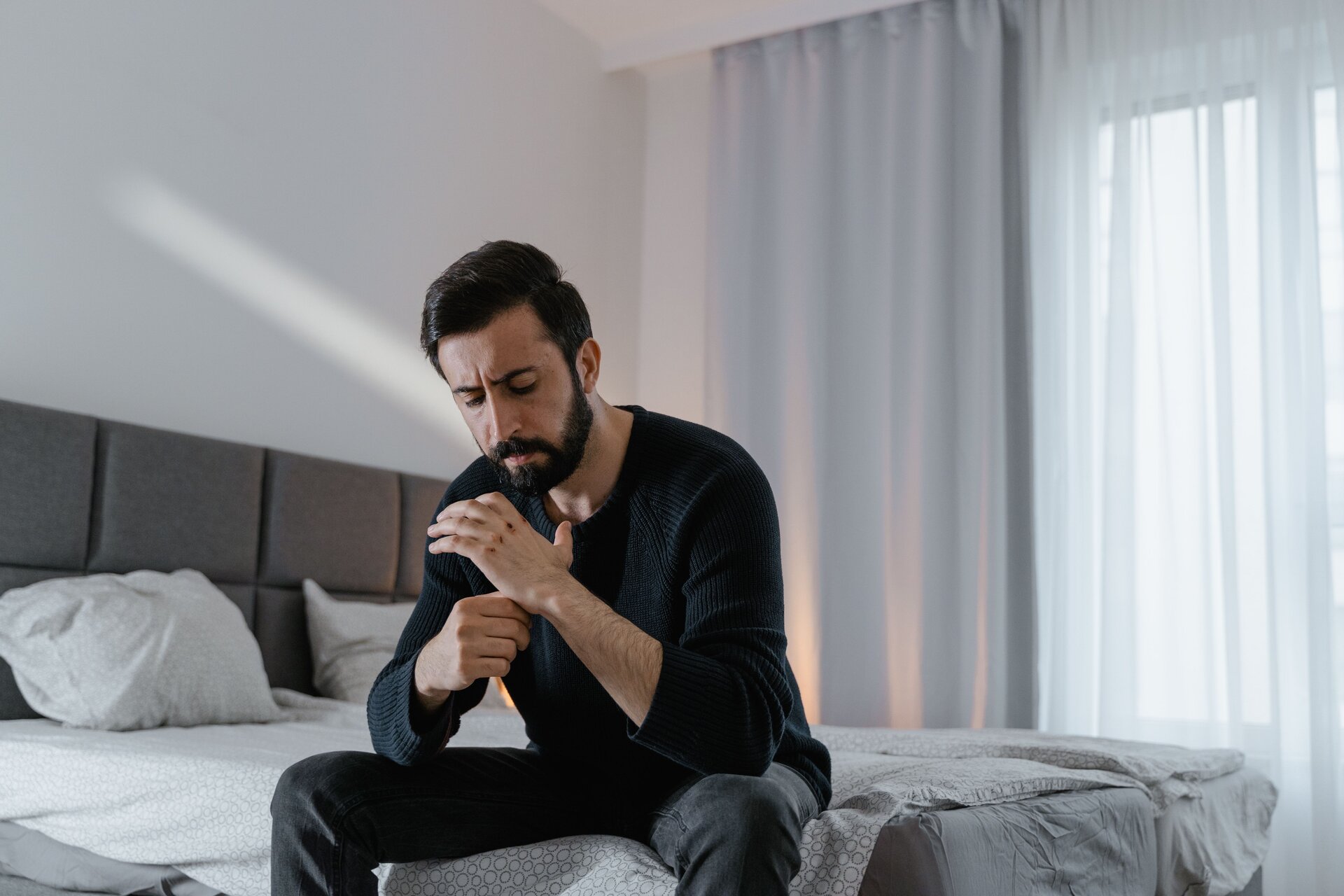 A man sitting on the edge of a bed while looking at and rubbing his hand