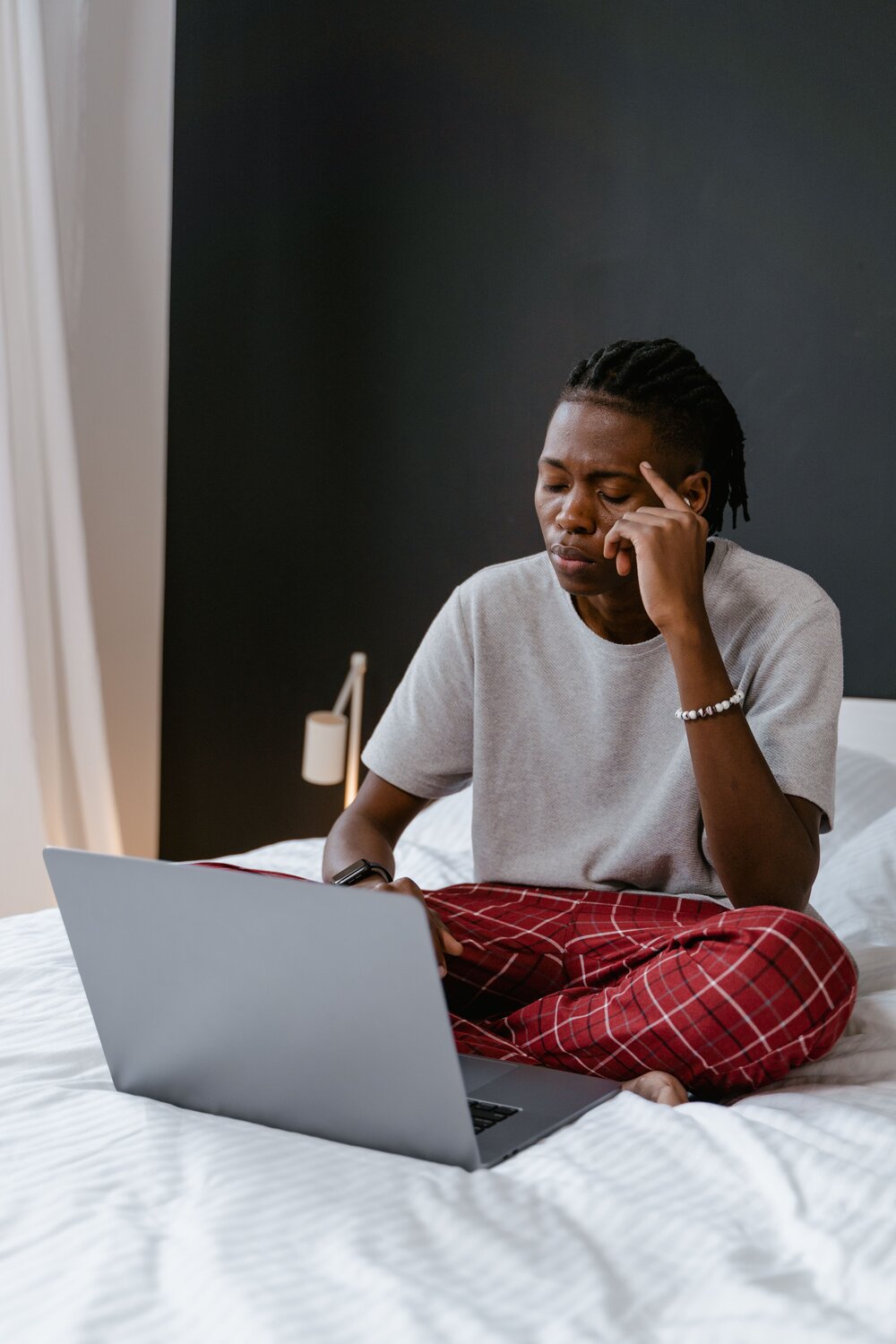 A man sitting on his bed with his laptop, while closing his eyes and rubbing his temple
