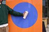 Guy in green holding a tan 24oz Owala tumbler in front of an art piece with orange and blue shapes.