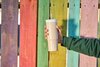 Person in a green sweater holding a tan 24oz Owala tumbler in front of a colorful fence.