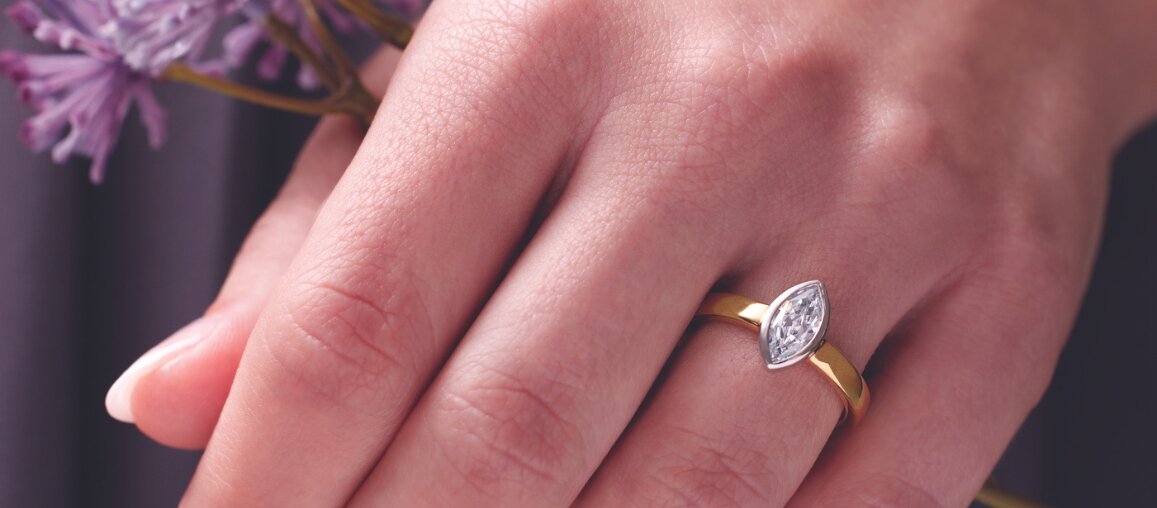 Minimalist marquise-cut diamond engagement ring in yellow gold worn on hand with soft floral background