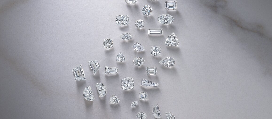 Loose diamonds in various cuts including round, emerald, oval, pear, cushion, and baguette arranged on a light stone background