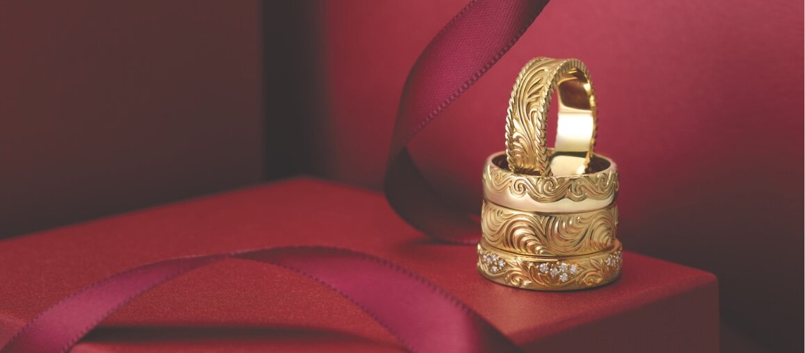 Stacked yellow gold wedding bands featuring intricate engraved scroll patterns and diamond accents, displayed on a red gift box with ribbon.
