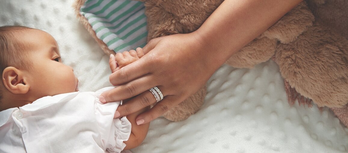 Newborn baby holding a mother’s hand as she wears stacked diamond wedding rings, symbolizing postpartum changes and ring fit after pregnancy