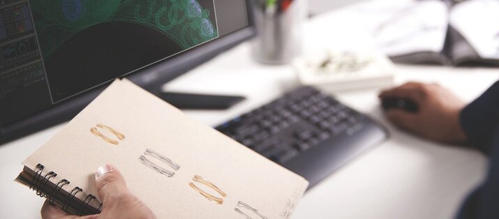 Jewelry designer sketching wedding bands in notebook with CAD ring model displayed on computer screen in Chicago studio