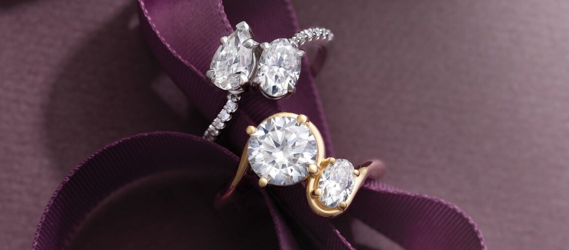 Three-stone diamond engagement rings with round and oval center stones and pear side diamonds on a plum ribbon background