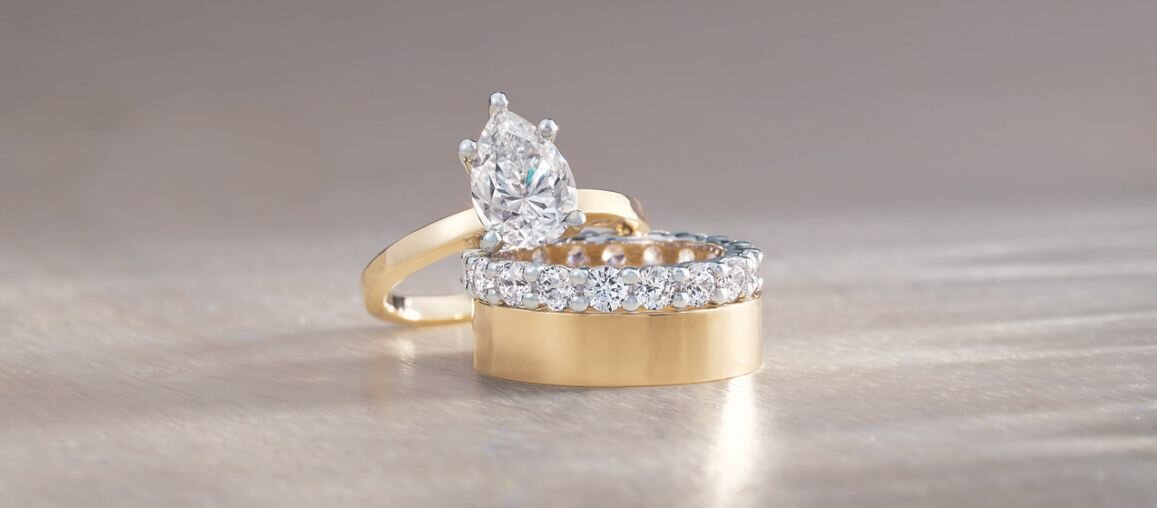 Stacked yellow gold wedding band paired with a diamond eternity band and a pear-cut diamond engagement ring, photographed on a neutral surface to highlight craftsmanship and sparkle.