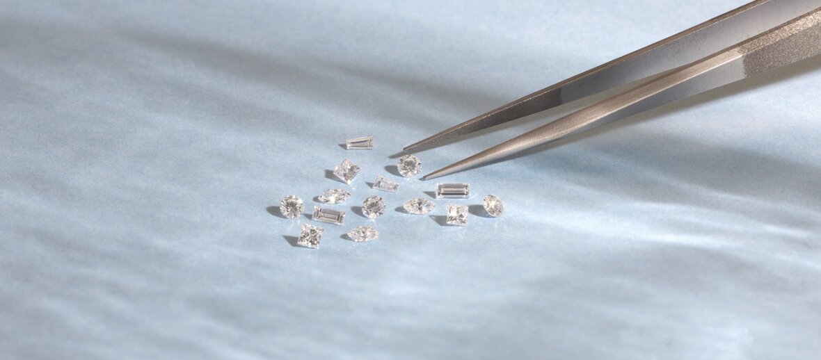 Close-up of loose diamonds in round, emerald, marquise, and baguette cuts being examined with jeweler tweezers on a light blue background