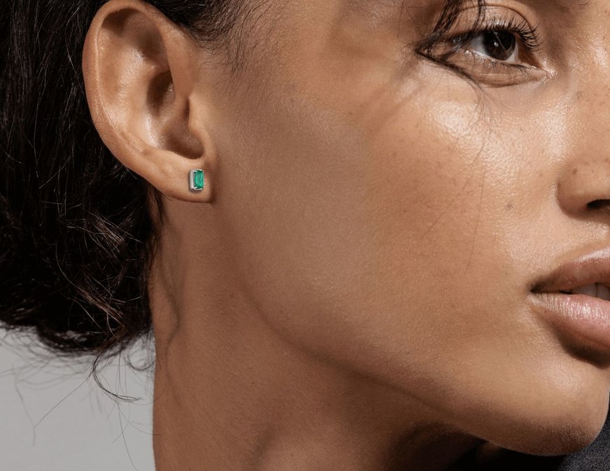 Image: A close-up view of a woman's earlobe adorned with elegant emerald stud earrings. The emerald gemstones glisten with a rich green hue.