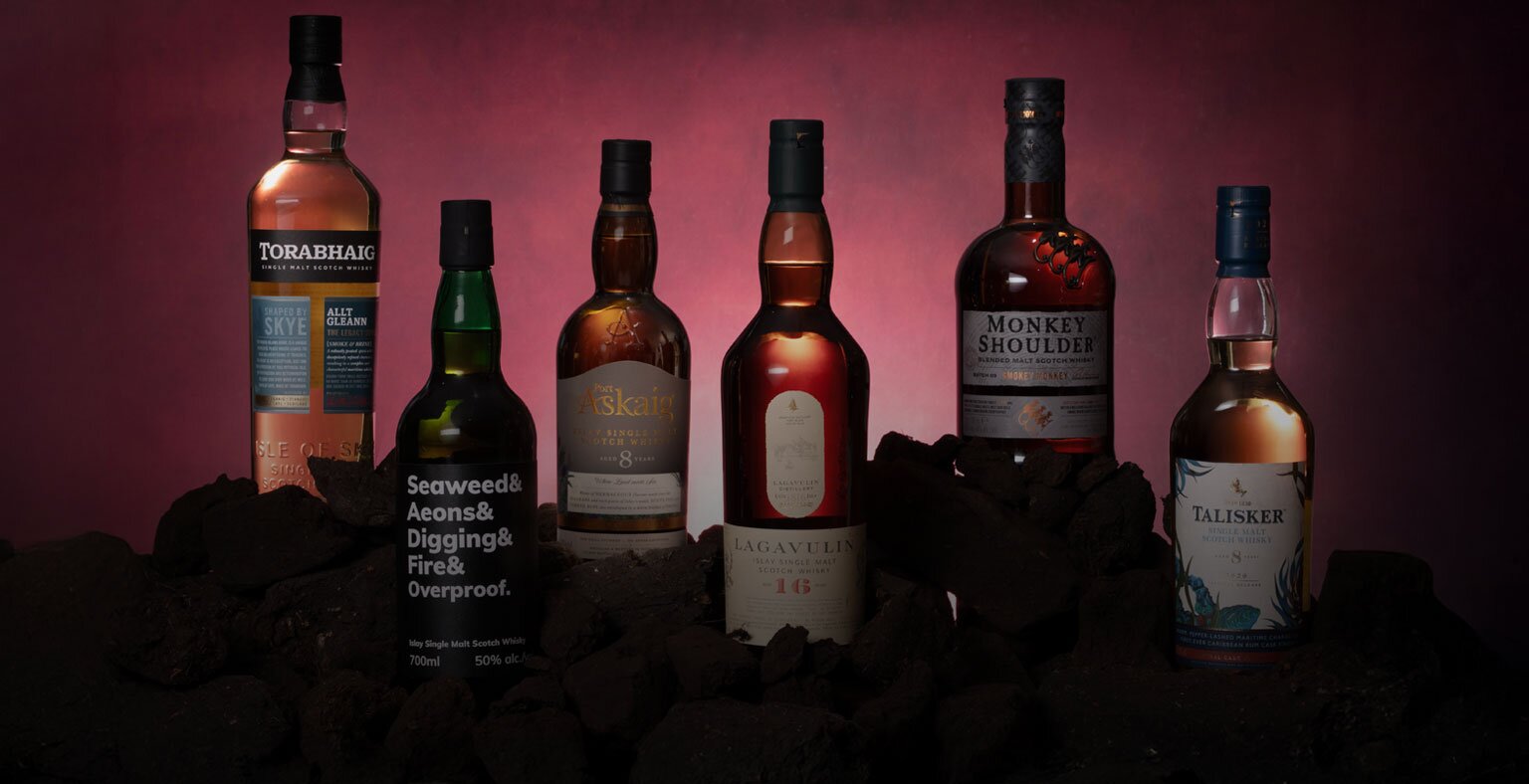 Six bottles of peated whisky against a red background surrounded by peat bricks