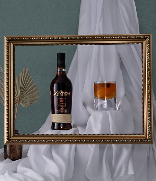 A bottle of Ron Zacapa next to a rum old fashioned cocktail behind a gold picture frame.